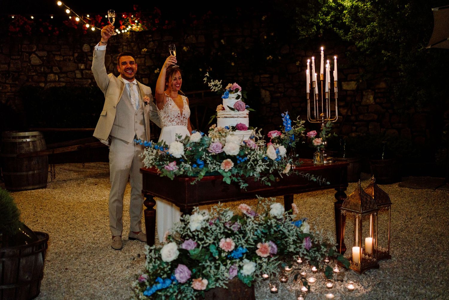 Bride and groom celebrating with sparklers beside floral wedding cake at candlelit outdoor evening reception.