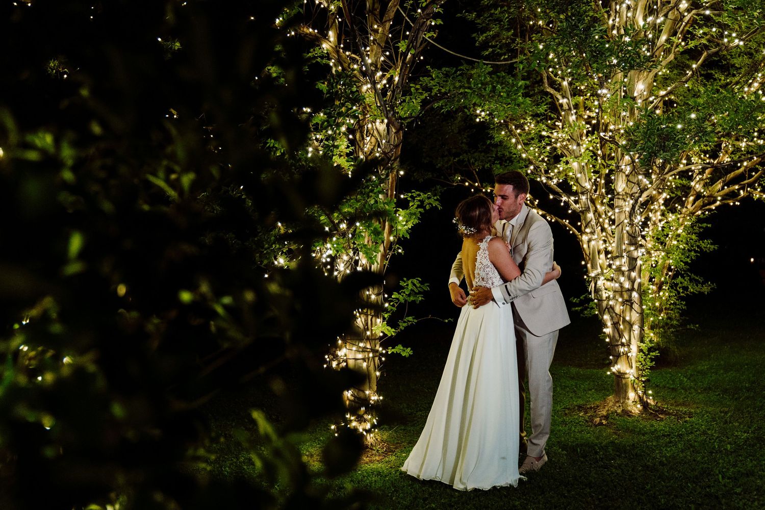Romantic bride and groom embracing under fairy light decorated trees in garden at night wedding.