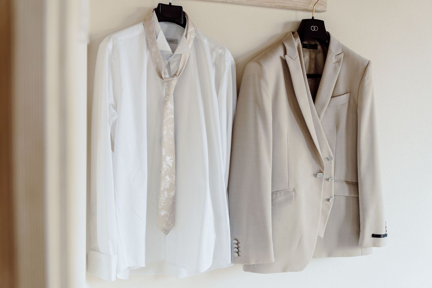 Groom's white dress shirt with tie and light beige wedding suit jacket hanging on wardrobe.