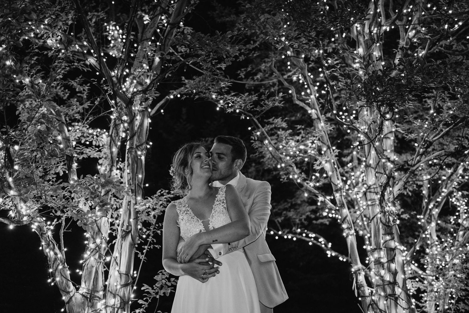 Black and white photo of bride and groom embracing under sparkling fairy lights in trees at night.