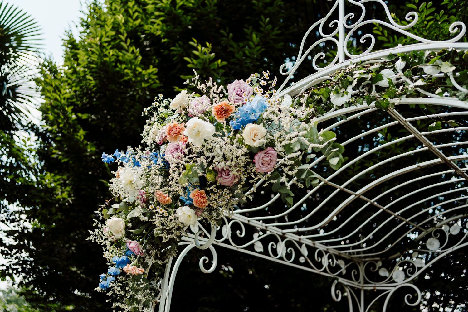 Elegant white wrought iron arch decorated with pink, blue and white floral arrangement for wedding ceremony.