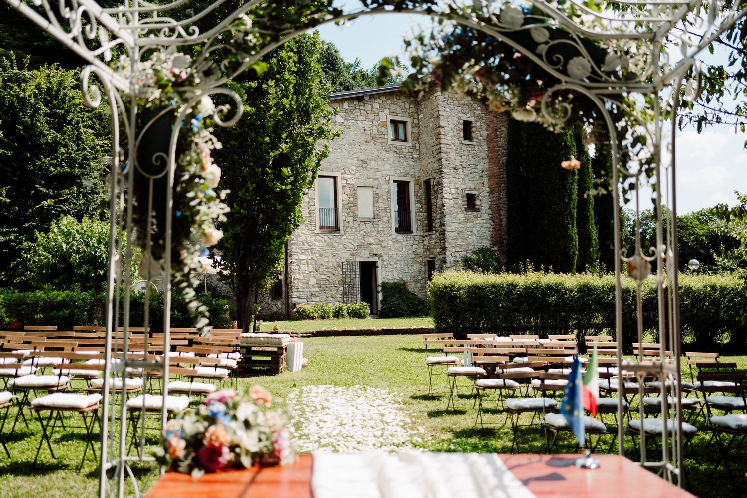 Outdoor wedding ceremony setup with white iron arch, wooden chairs and stone villa in background.