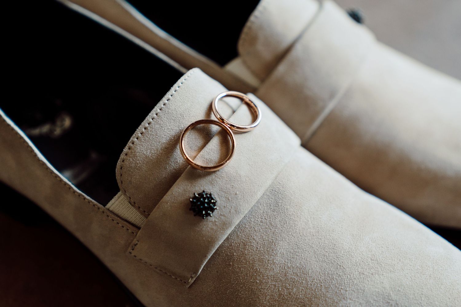Gold wedding rings resting on beige suede loafers with a decorative button detail.