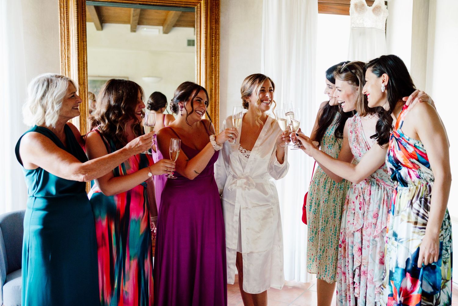 Bride in white robe toasting champagne with colorful-dressed bridesmaids before the wedding.
