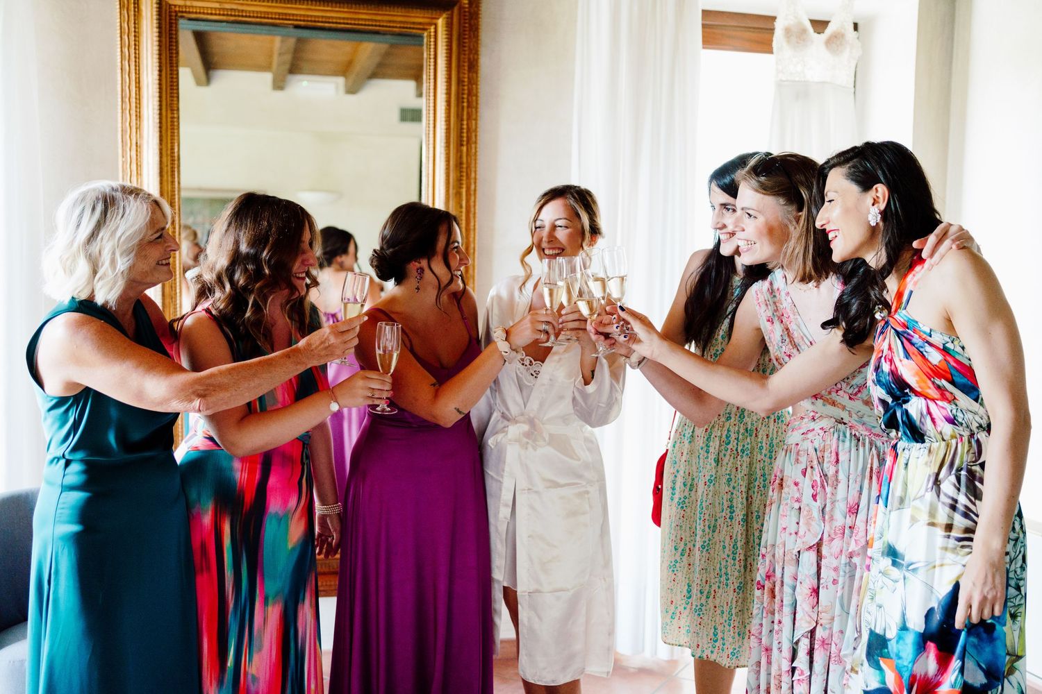 Joyful bride and bridesmaids clinking champagne glasses in vibrant dresses during bridal prep.