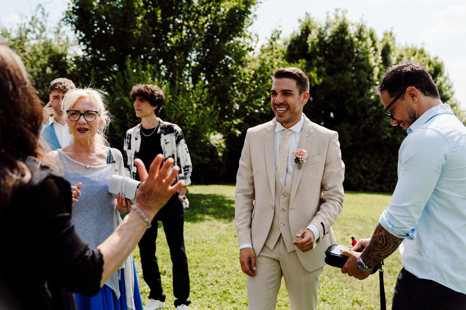 Smiling groom in beige suit greeting guests outdoors on his wedding day.