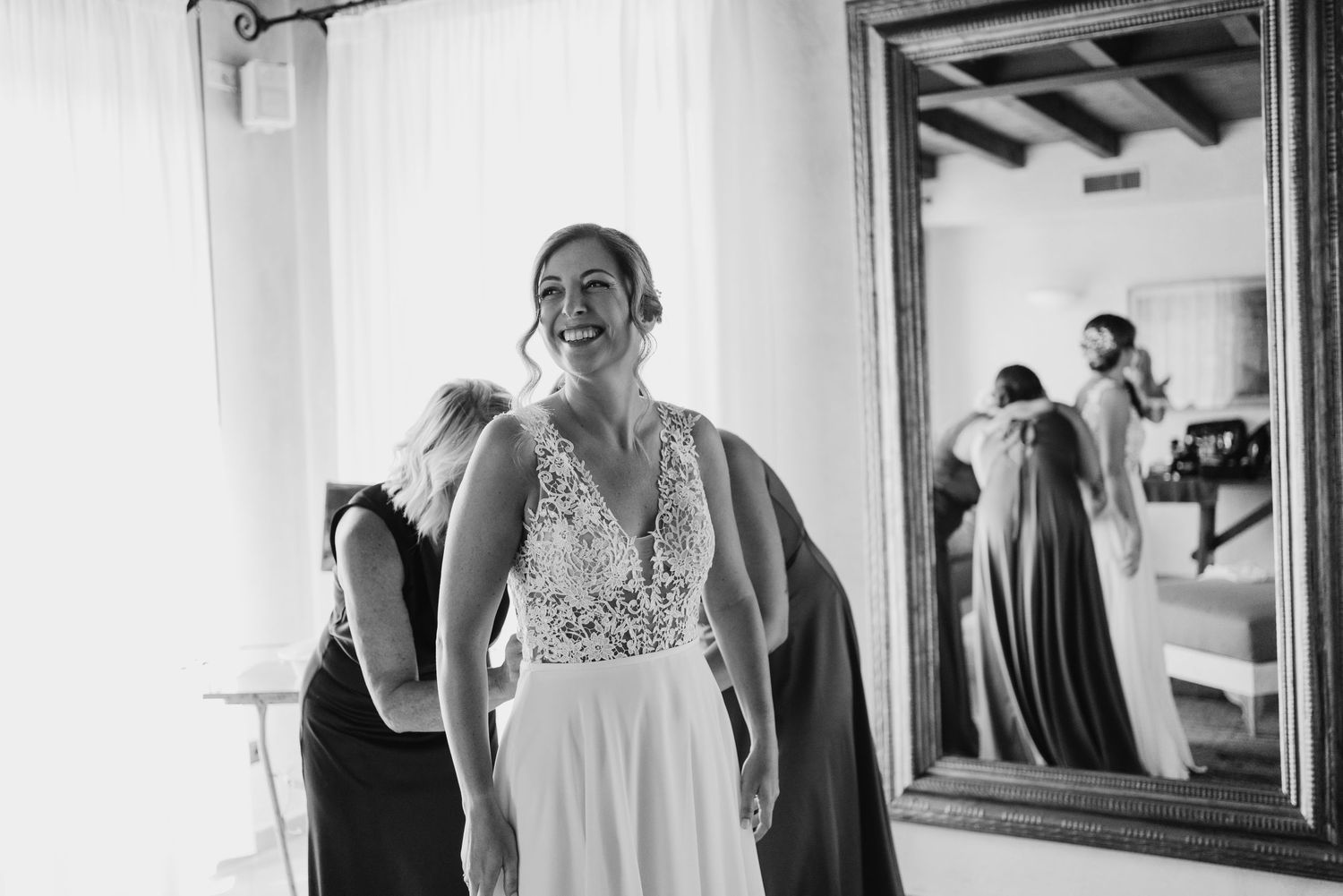 Bride laughing in white lace wedding gown while being helped by bridesmaids in black and white photo.