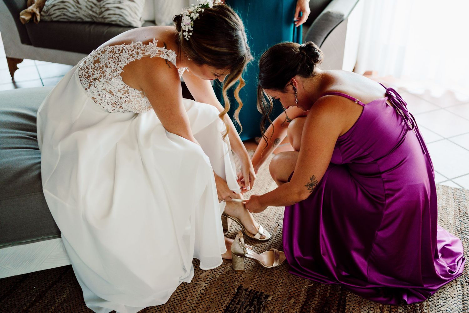 Bridesmaid in purple dress helping bride put on wedding shoes before ceremony.