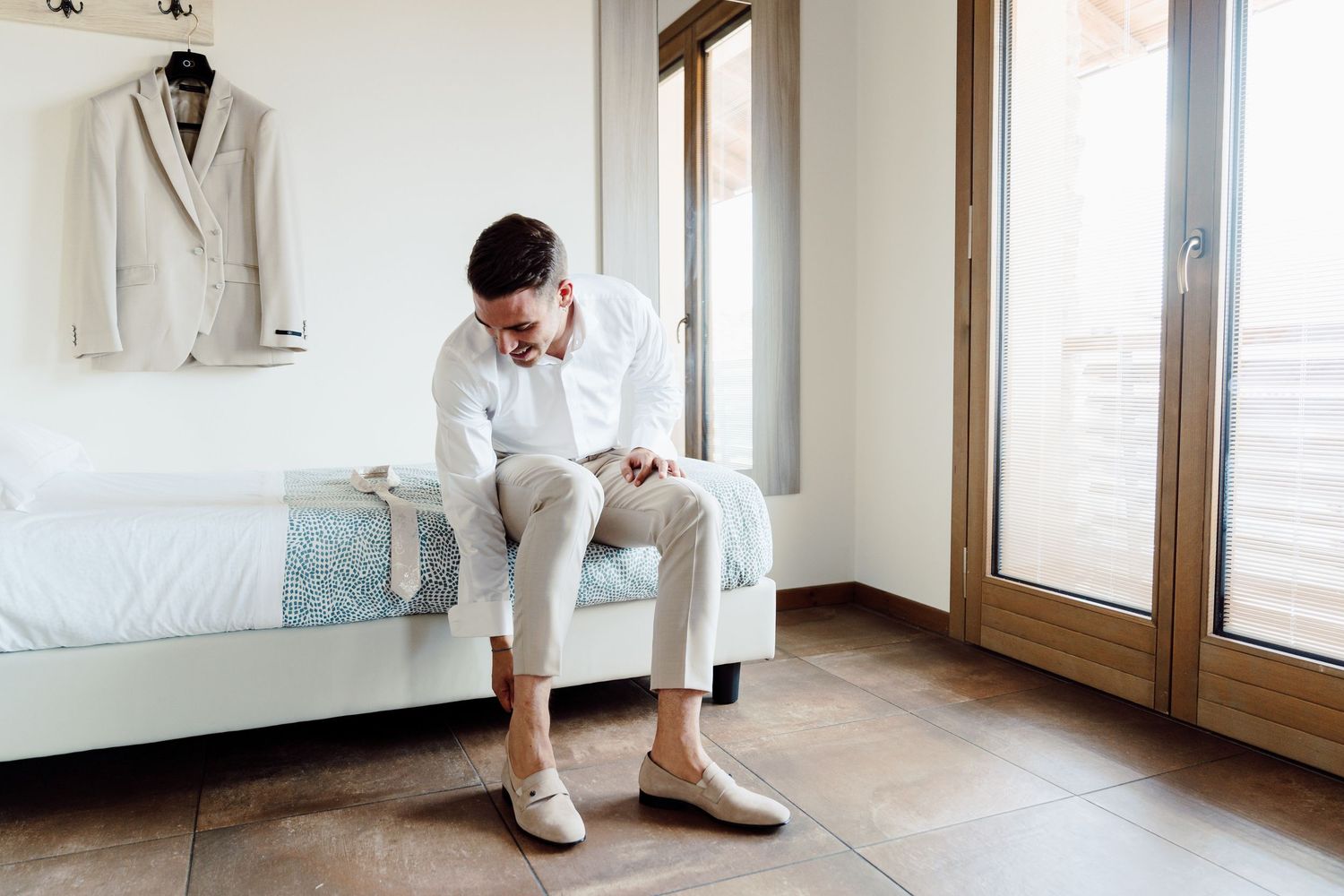 Groom in white shirt and beige trousers sitting on bed putting on shoes in bright hotel room.