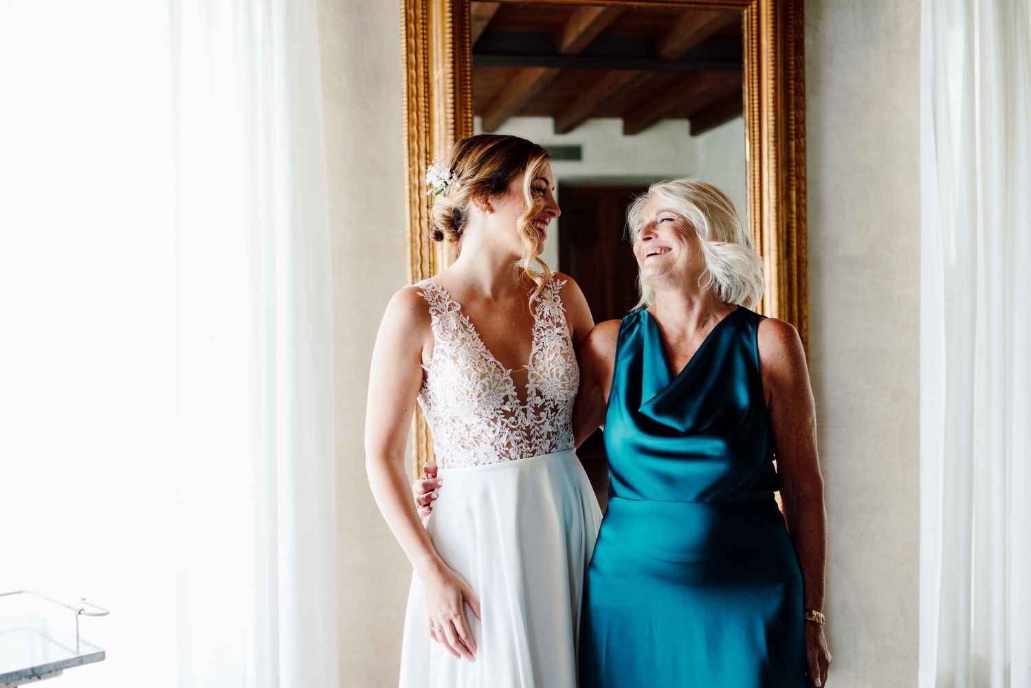 Bride in lace wedding gown sharing a joyful moment with older woman in teal dress by mirror.