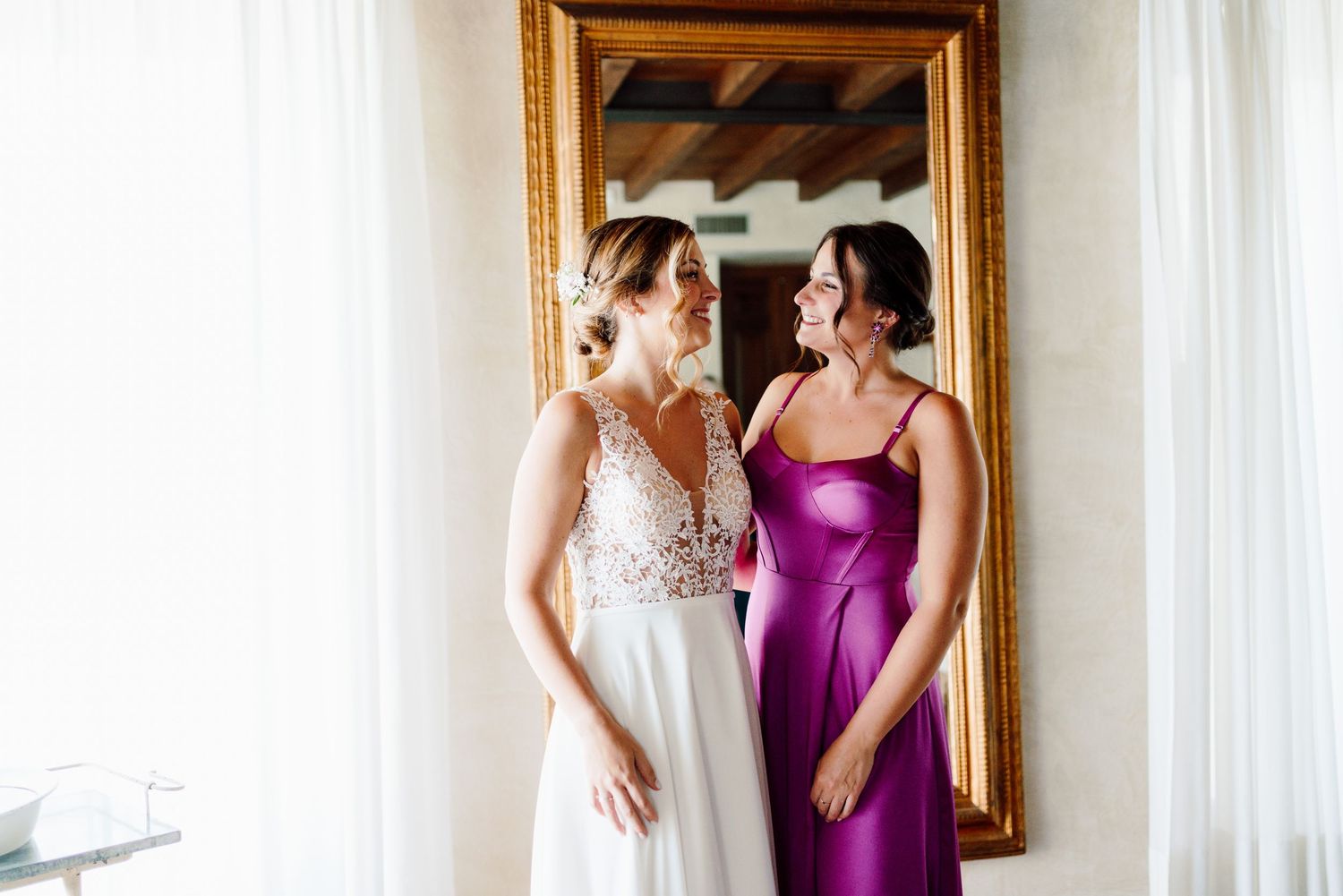 Bride in V-neck lace wedding dress smiling with bridesmaid in purple gown near gold mirror.