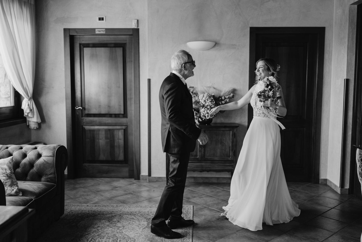 Black and white photo of bride receiving bouquet from suited man in rustic interior.
