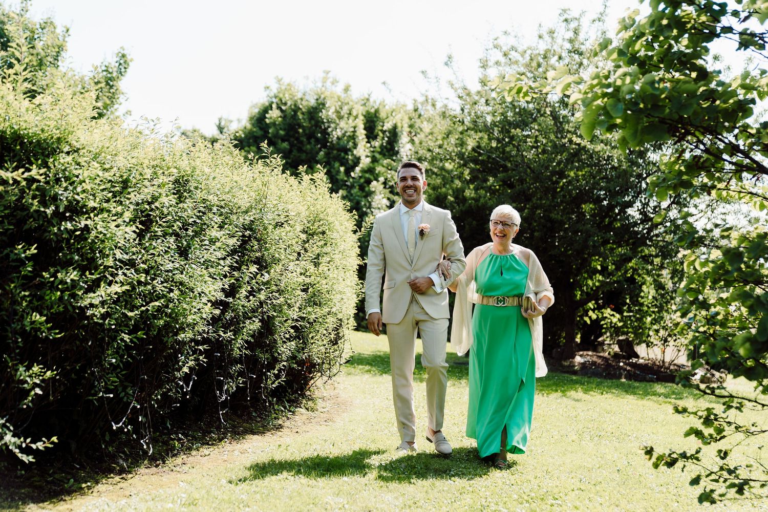 Groom in beige suit walking with elderly woman in green dress through garden path.