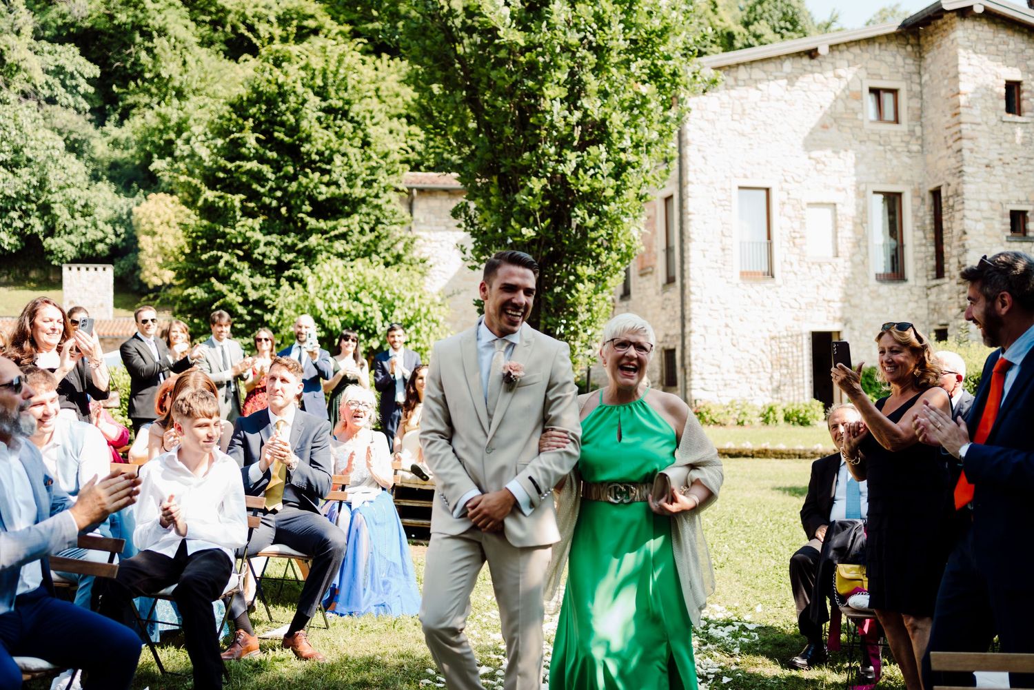 Groom in linen suit escorting woman in green dress down outdoor wedding aisle with guests.
