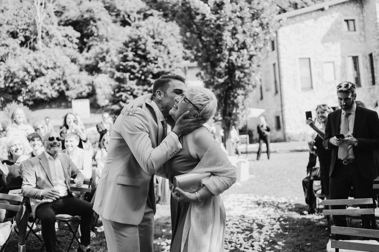 Couple sharing first kiss at outdoor wedding ceremony with guests seated in background.