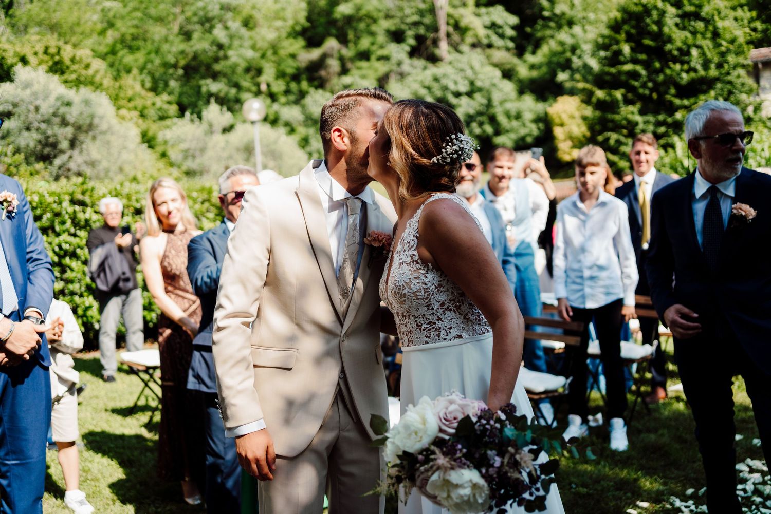Newlyweds sharing romantic kiss outdoors while wedding guests celebrate around them.