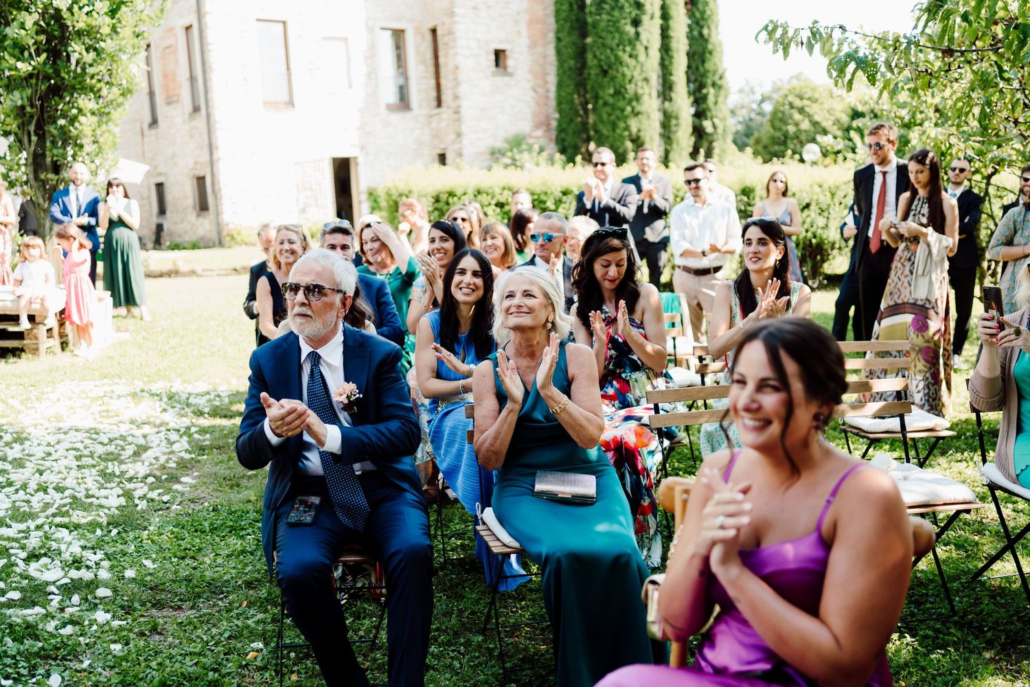 Wedding guests clapping and smiling during outdoor ceremony on sunny day at historic venue.