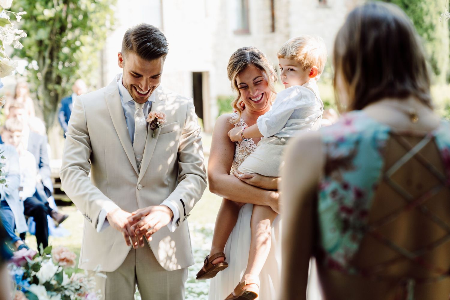 Smiling groom in beige suit and bride holding toddler greeted by guests after ceremony.