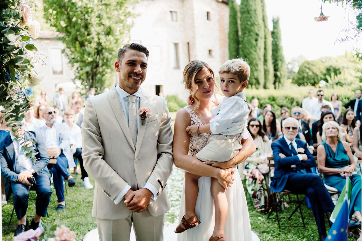 Groom in tan suit and bride carrying young child walking down wedding aisle together.