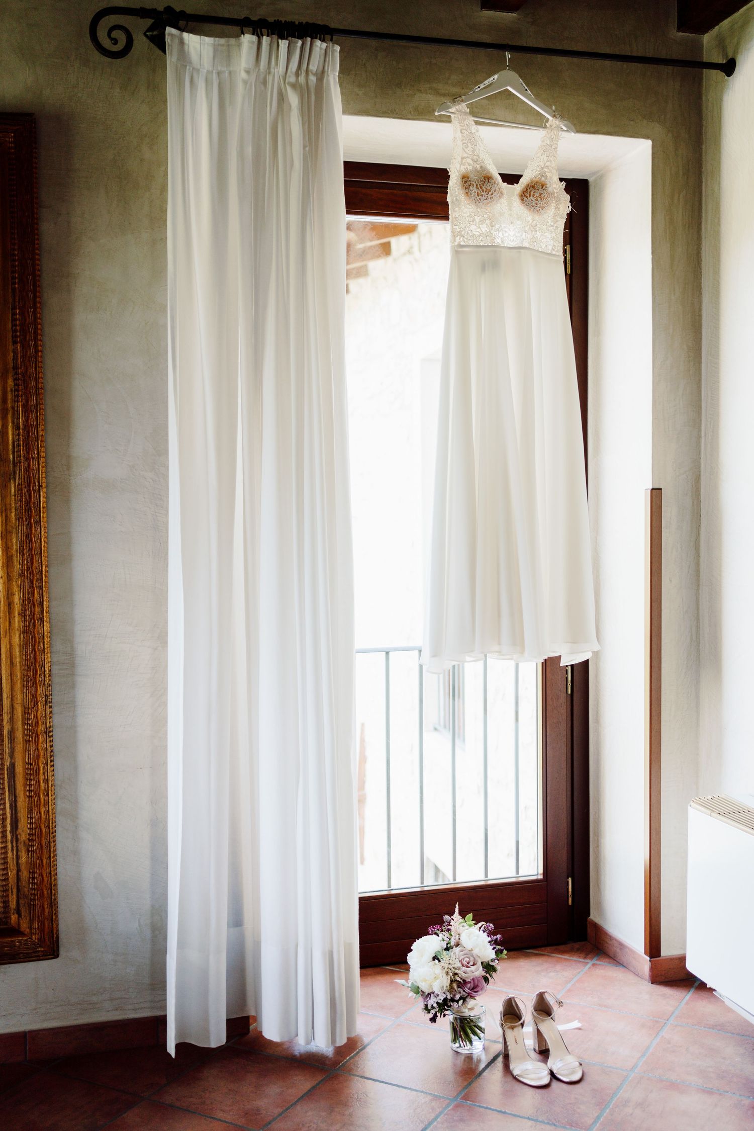 Elegant white wedding dress hanging by window with bridal bouquet and heels below.