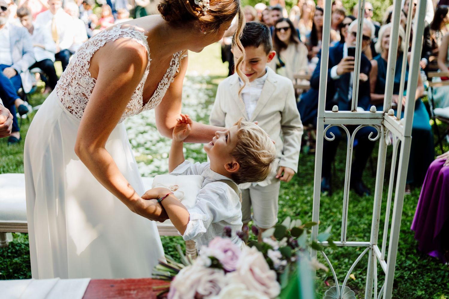 Bride in lace gown bending down to interact with young child during outdoor wedding ceremony.