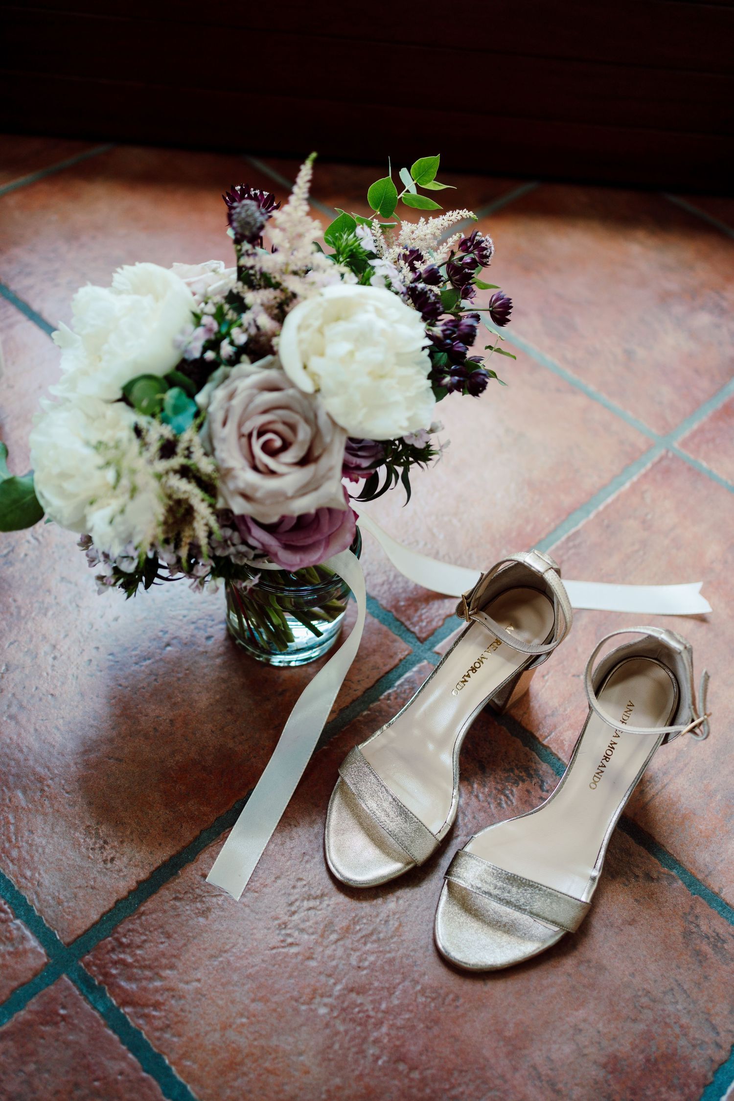 Elegant bridal gold sandals beside a bouquet of white and purple flowers on terracotta tiles.