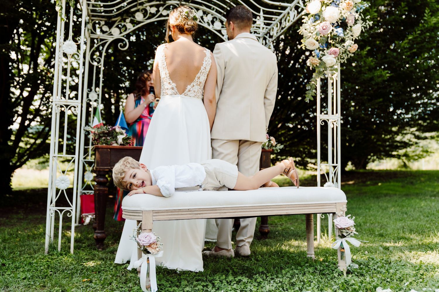 Young boy lies on a bench behind bride and groom during an outdoor wedding ceremony.