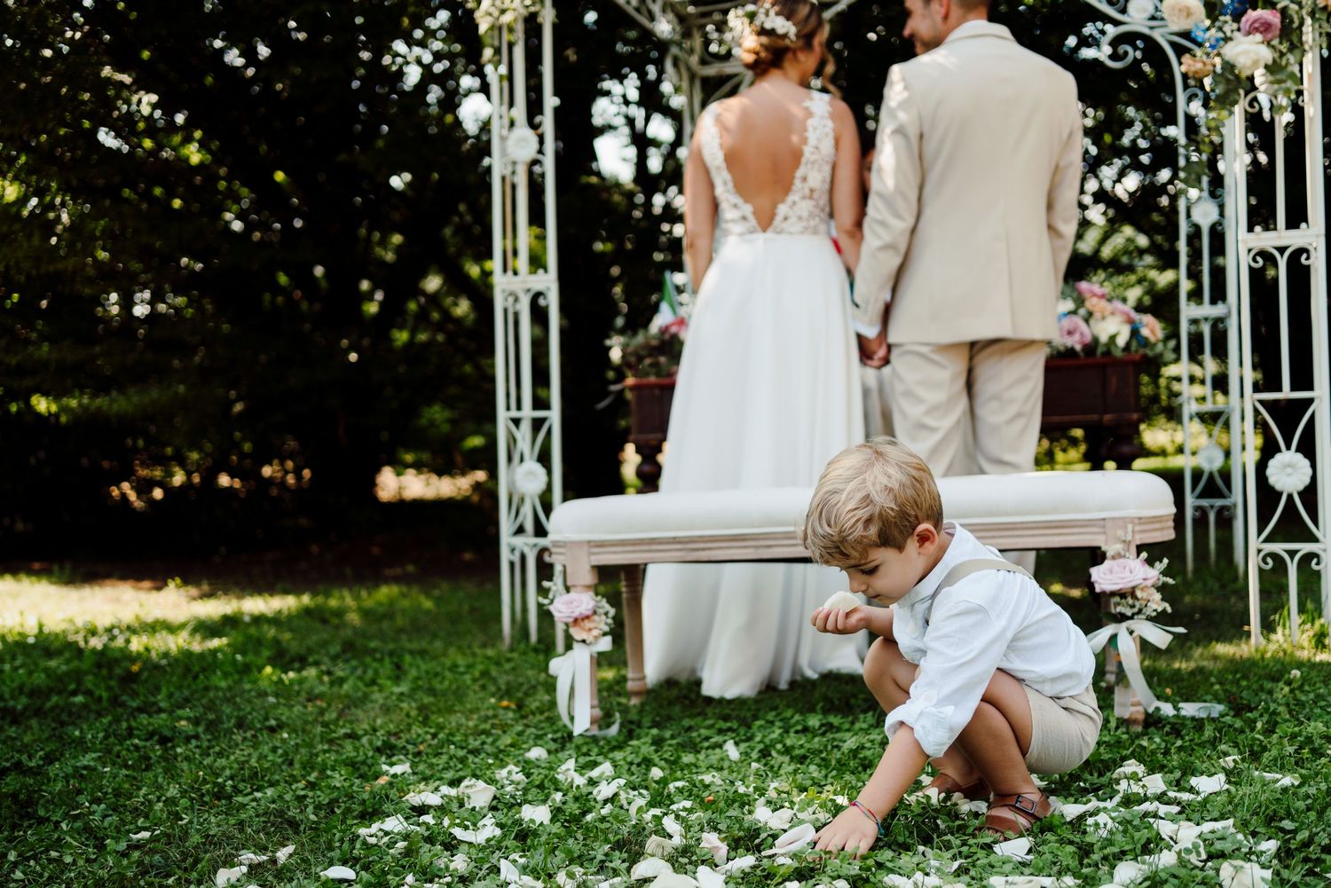 Young ring bearer picks up flower petals from the ground during an outdoor wedding ceremony.