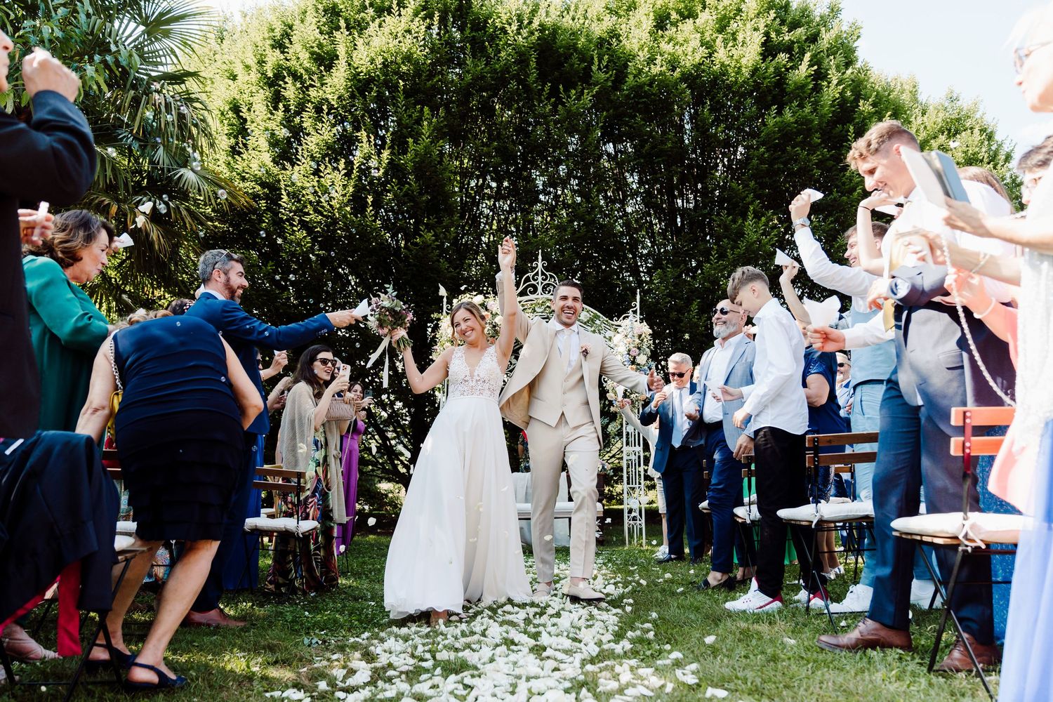 Bride and groom walk joyfully through a petal-lined aisle surrounded by cheering wedding guests.