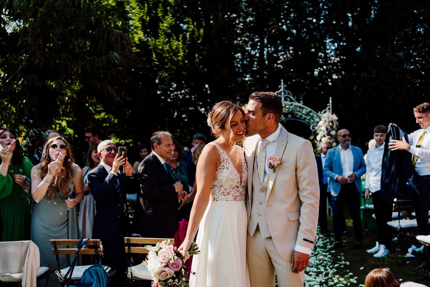 Bride and groom share a kiss at outdoor wedding ceremony while guests applaud and celebrate around them.