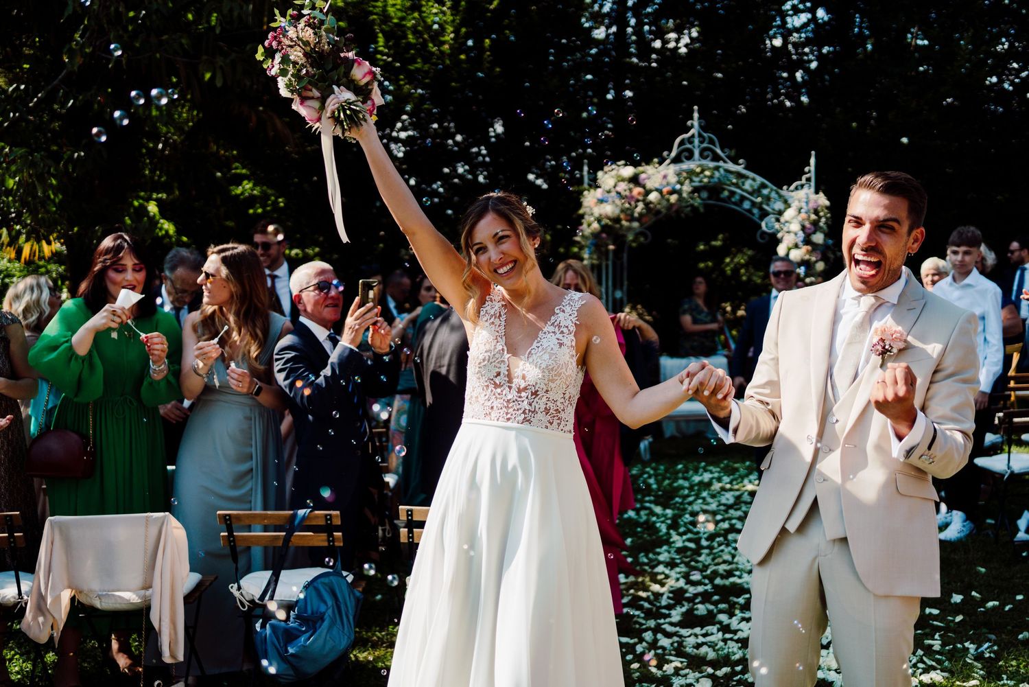 Joyful bride raises bouquet triumphantly as groom smiles beside her after outdoor wedding ceremony.