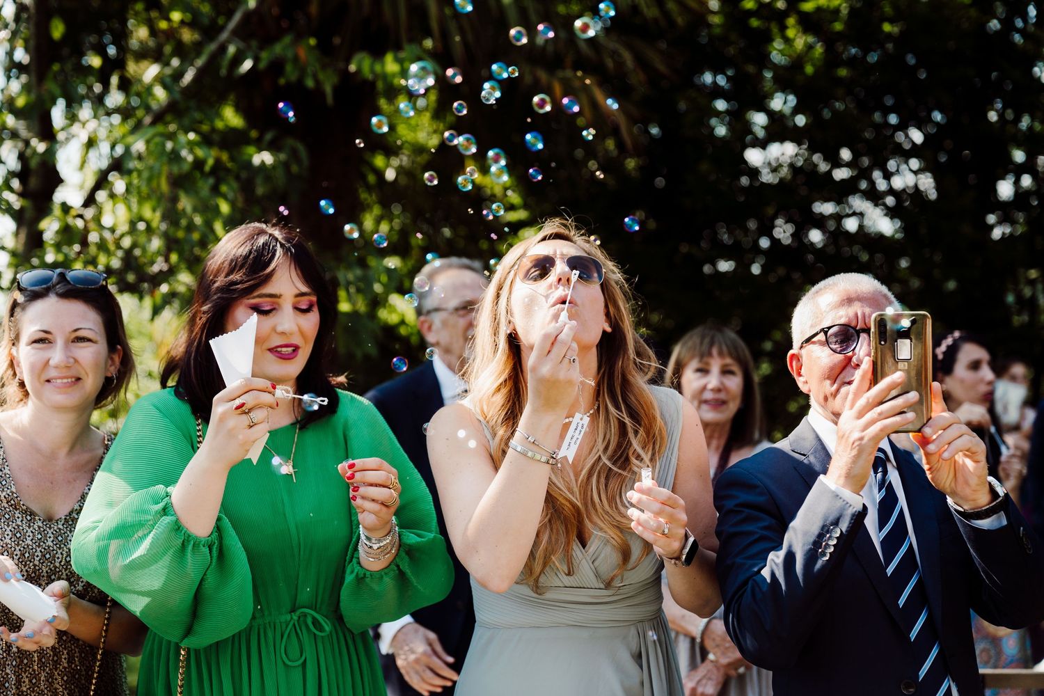 Wedding guests blow bubbles in celebration outdoors, woman in green dress and man with red glasses.