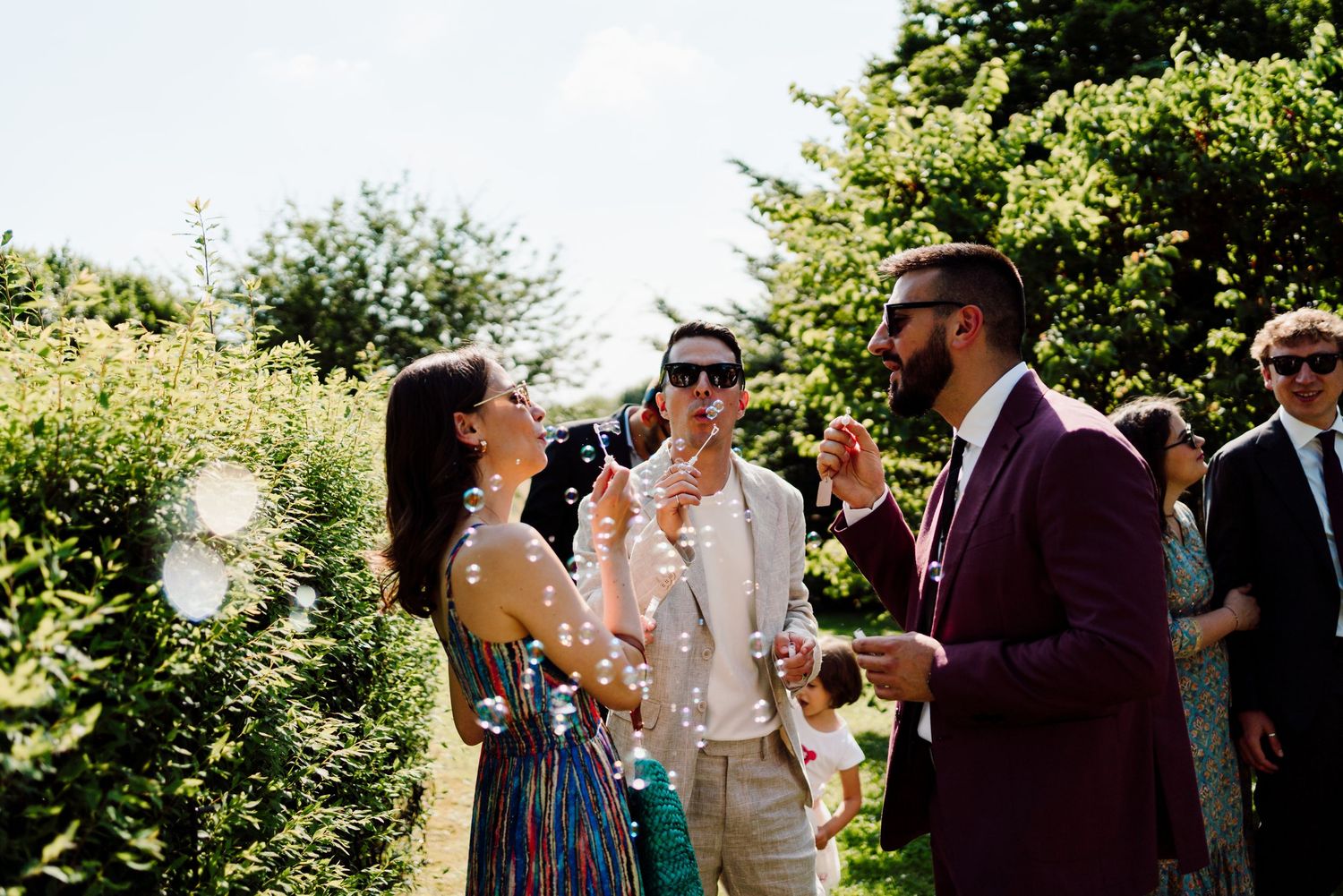 Wedding guests blow bubbles outdoors, man in burgundy suit and woman in colorful dress celebrating.