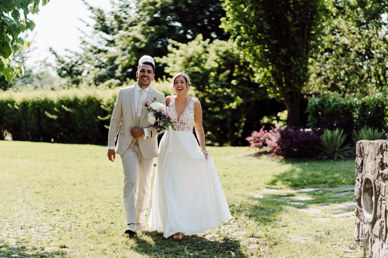 Bride and groom walk hand in hand through sunny garden after outdoor wedding ceremony.