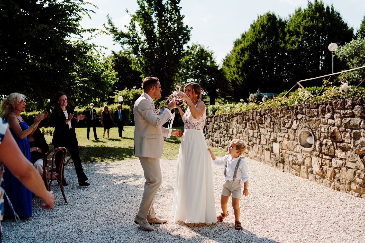 Bride and groom toast champagne glasses outdoors while small child stands nearby at wedding celebration.
