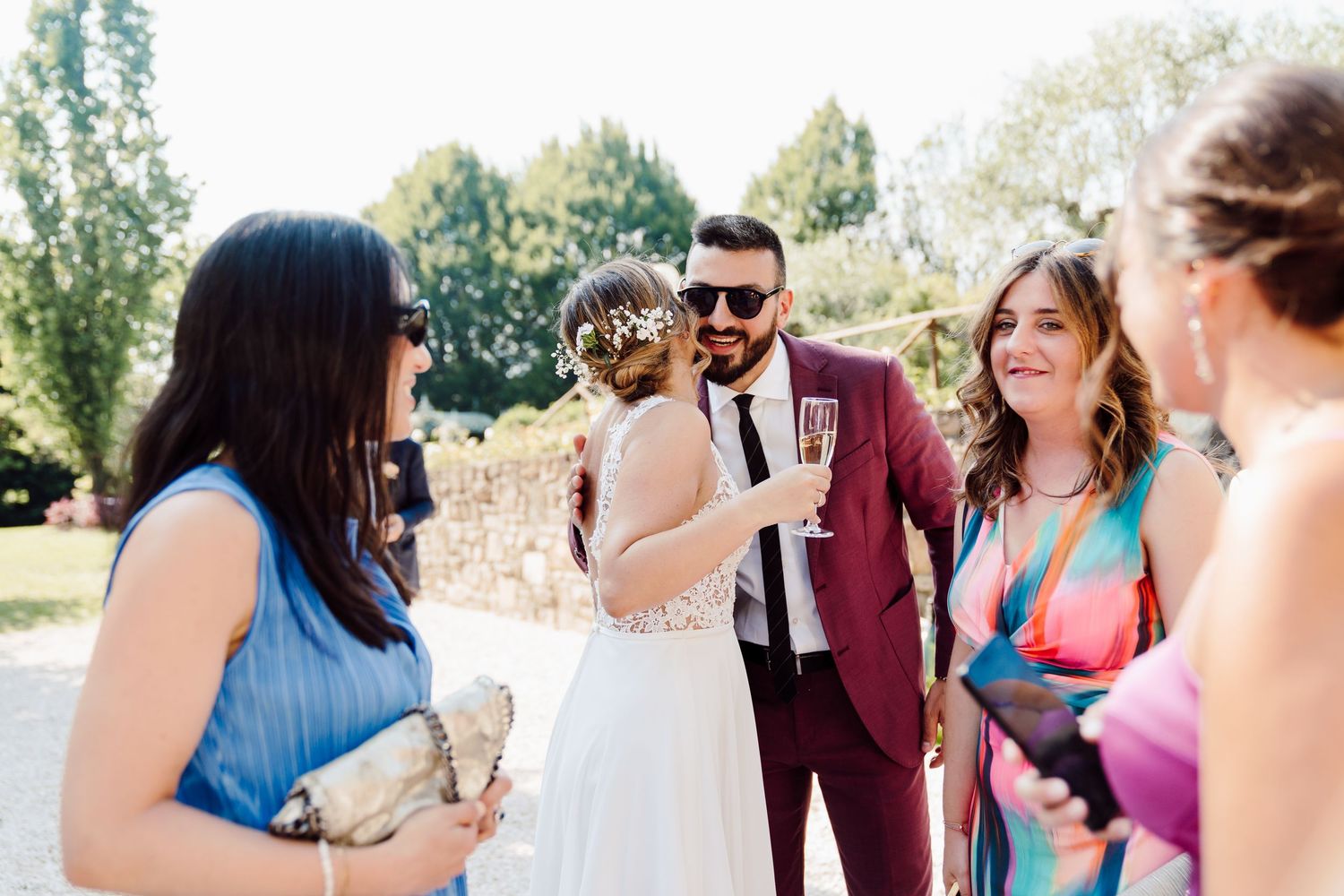 Bride in lace gown chats with guests including man in burgundy suit at sunny outdoor wedding reception.