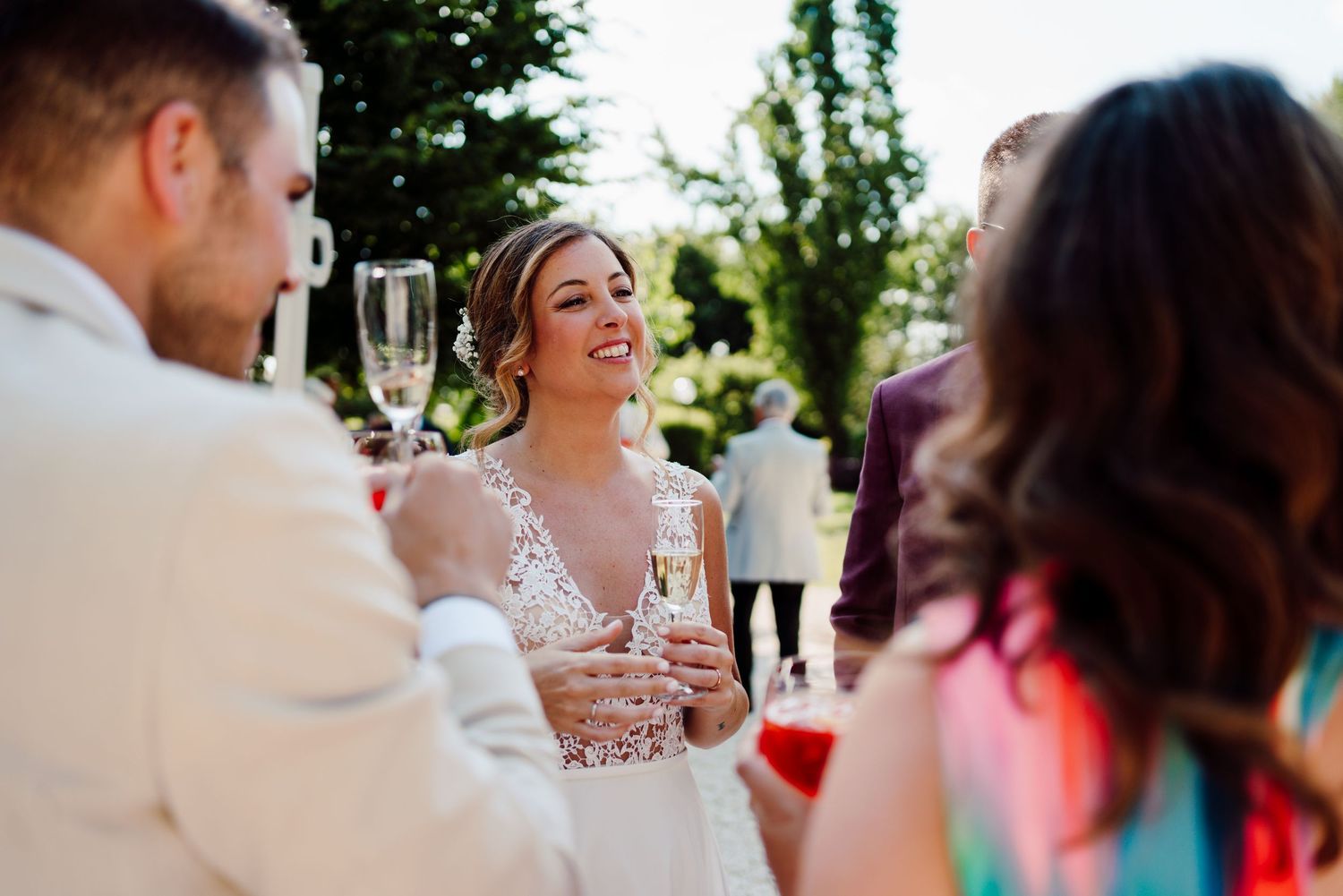 Smiling bride in lace wedding dress toasting champagne with guests at outdoor wedding reception.