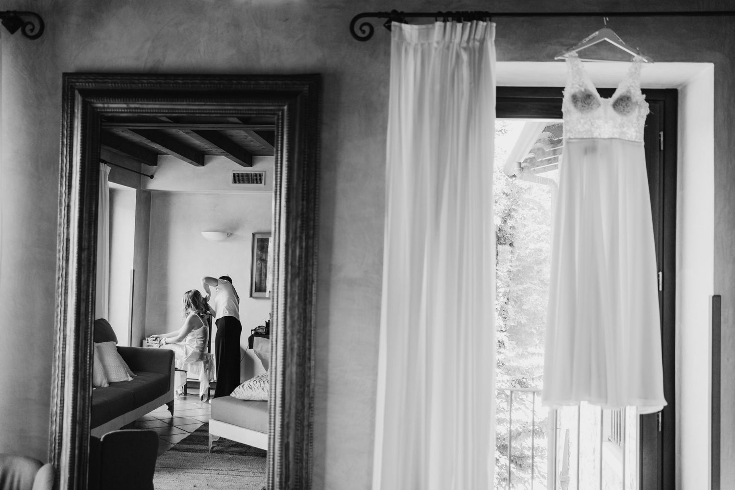 Black and white photo of wedding dress hanging by window with bride getting ready in mirror.