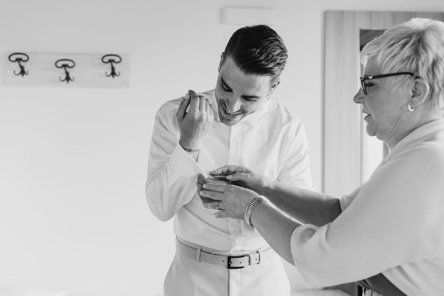 Black and white photo of groom getting cufflinks fastened by older woman before wedding.