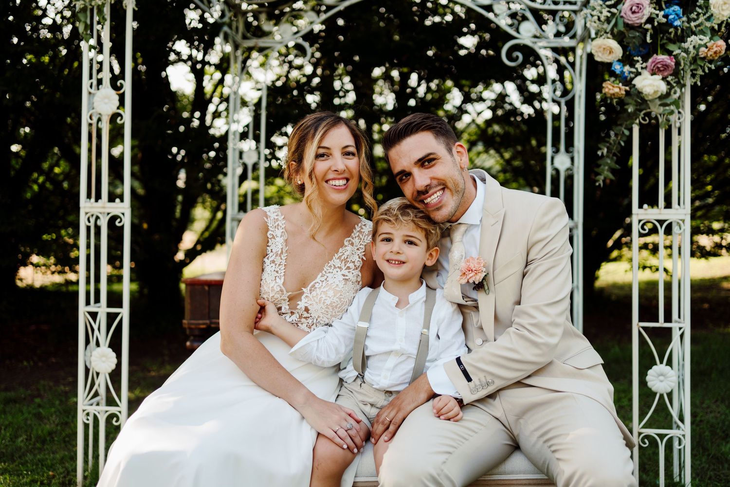 Bride and groom sitting under white floral arch with young boy in white suit at wedding.