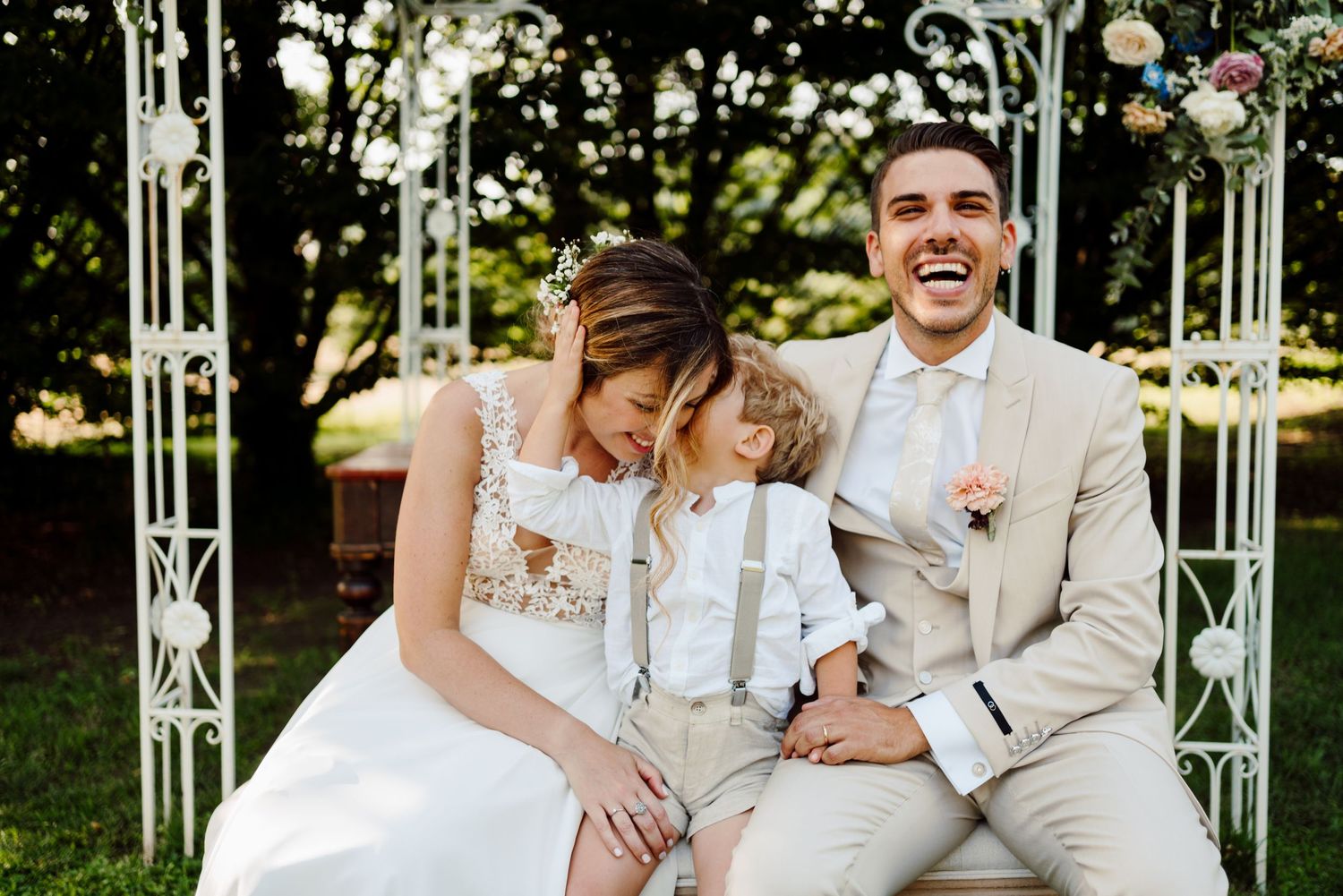 Laughing bride and groom seated under white wedding arch with young child between them.