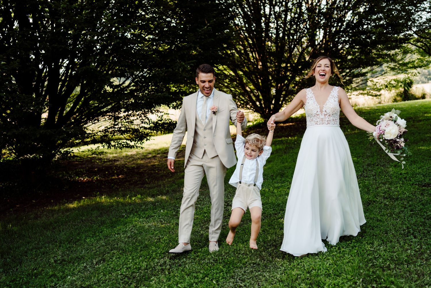 Joyful bride and groom in beige suit swinging young boy between them in green garden.