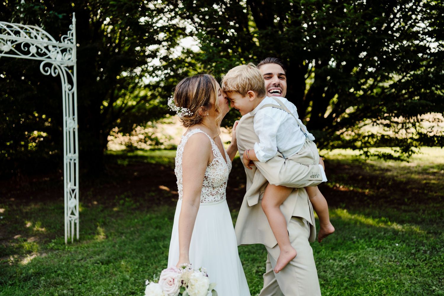 Groom laughing as young boy kisses his cheek while bride holds bouquet in garden.