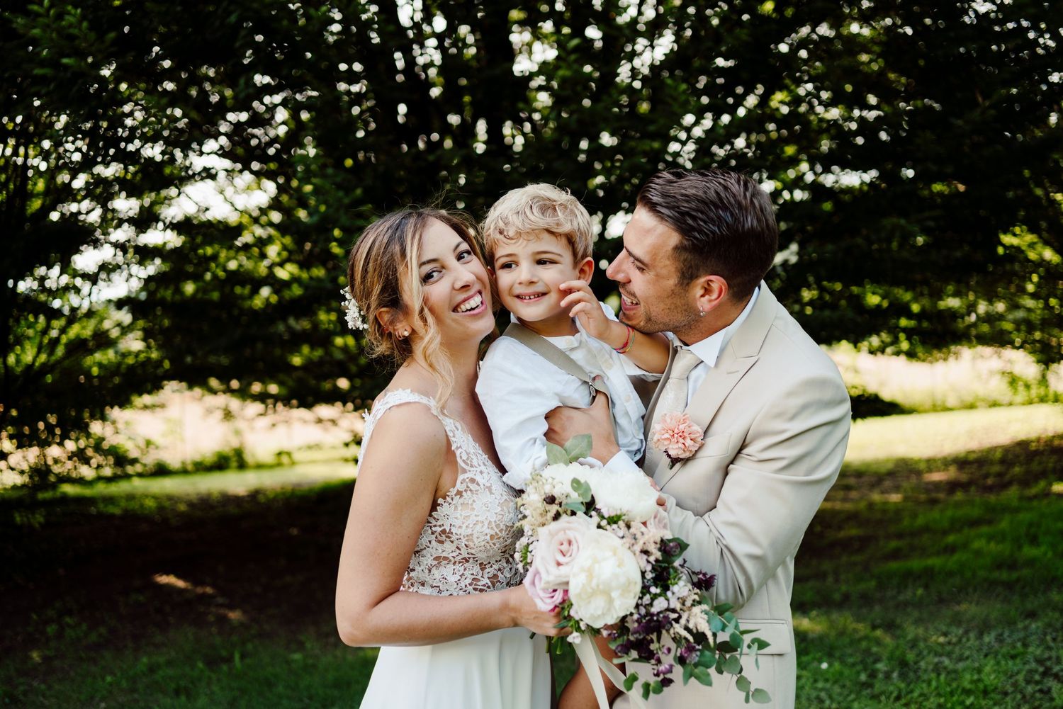 Bride and groom holding young blond boy while posing with bouquet in lush garden setting.