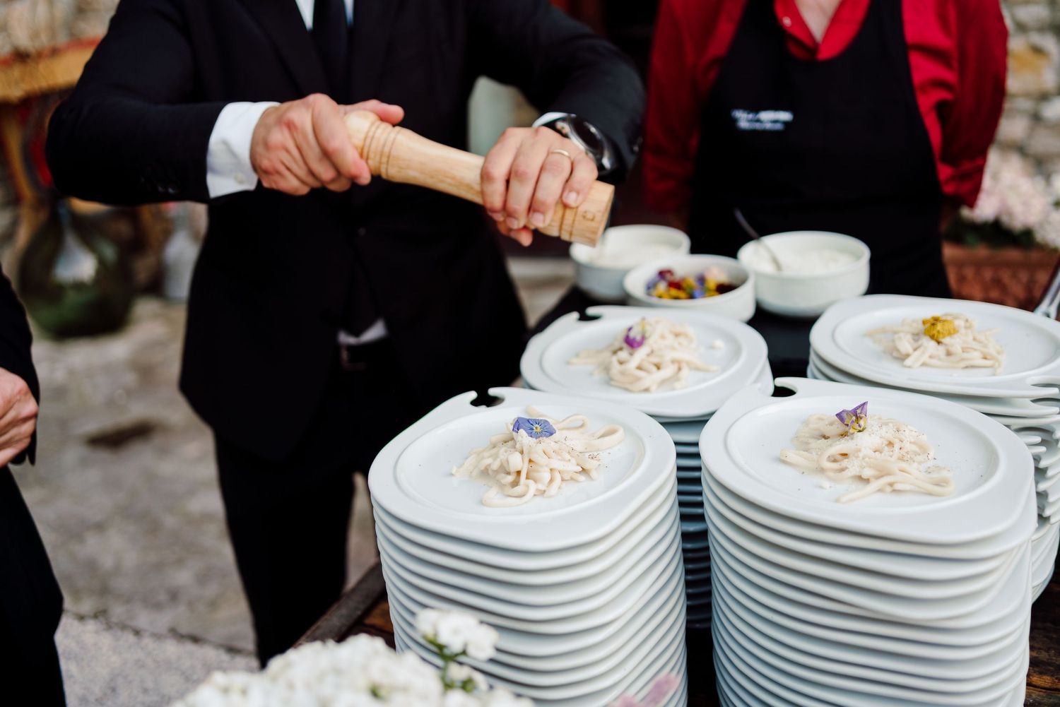 Catering staff plating elegant pasta dishes on white plates at an outdoor wedding reception.