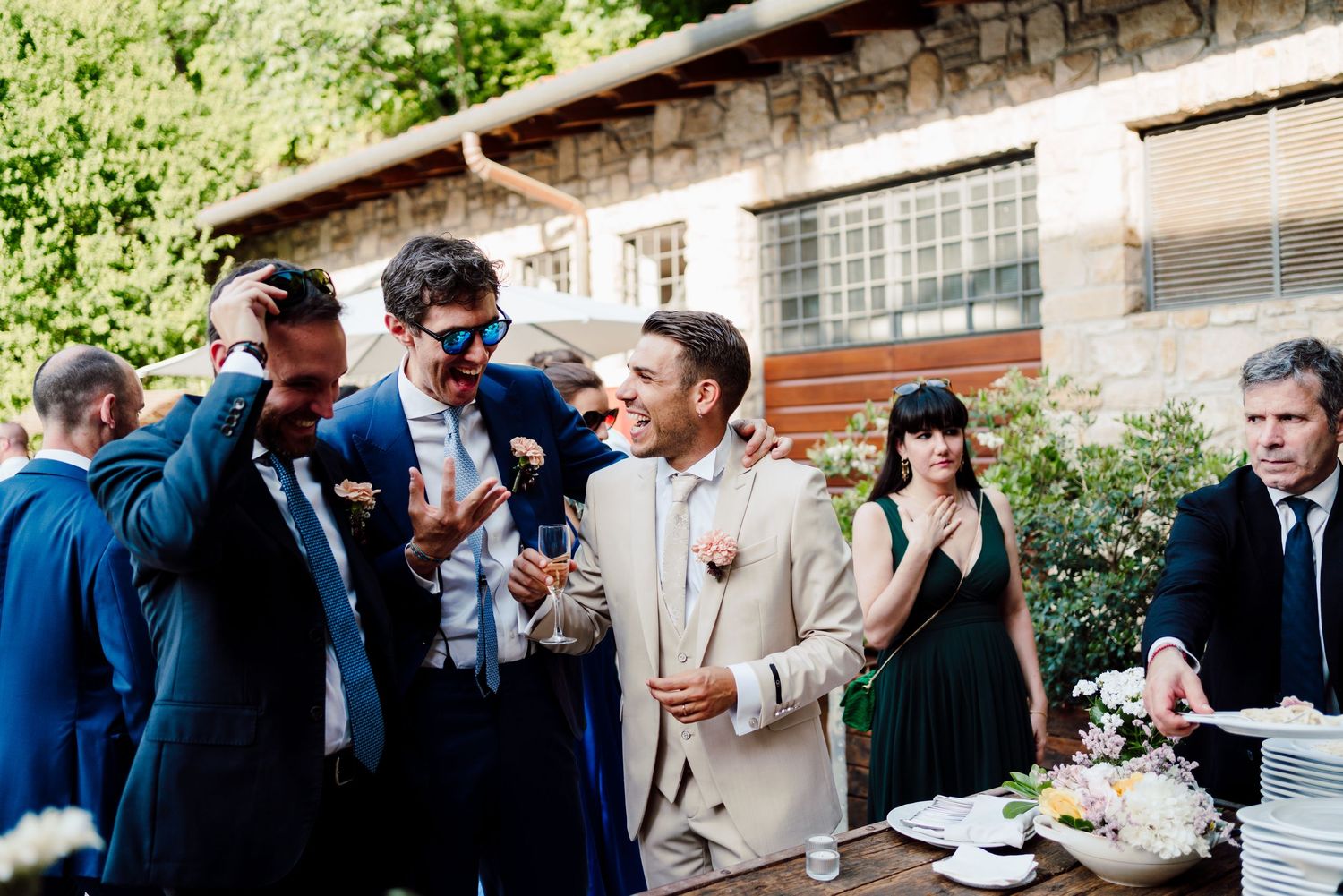 Groomsmen in navy suits laughing and celebrating at an outdoor wedding cocktail hour.