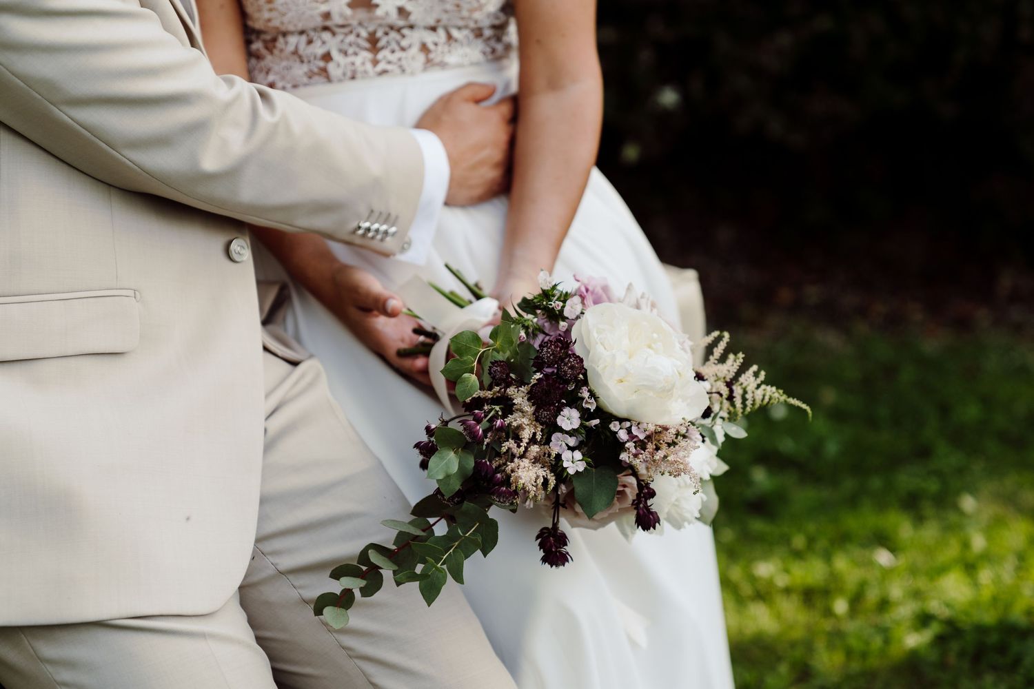 Close-up of bride and groom holding a beautiful bouquet with white peonies and burgundy blooms.