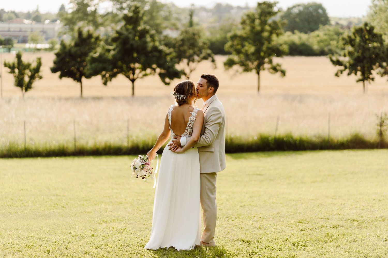 Newlyweds embracing and kissing on a green lawn with a scenic countryside backdrop.