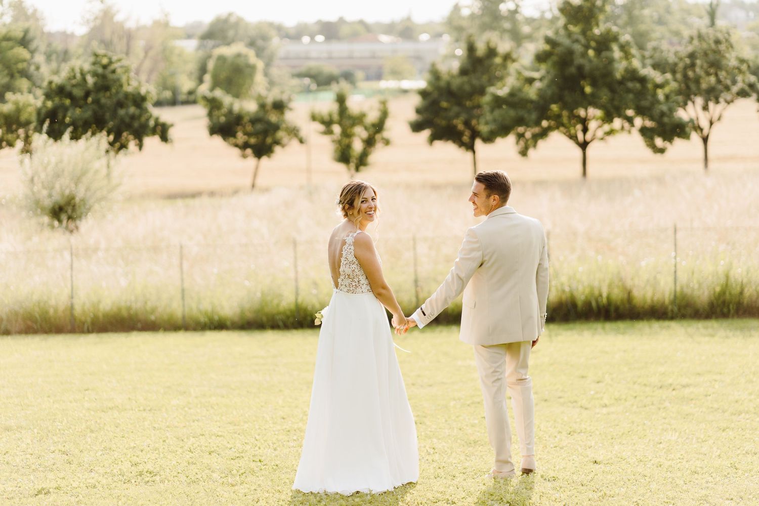 Bride and groom holding hands and gazing at each other in a sunlit field on their wedding day.