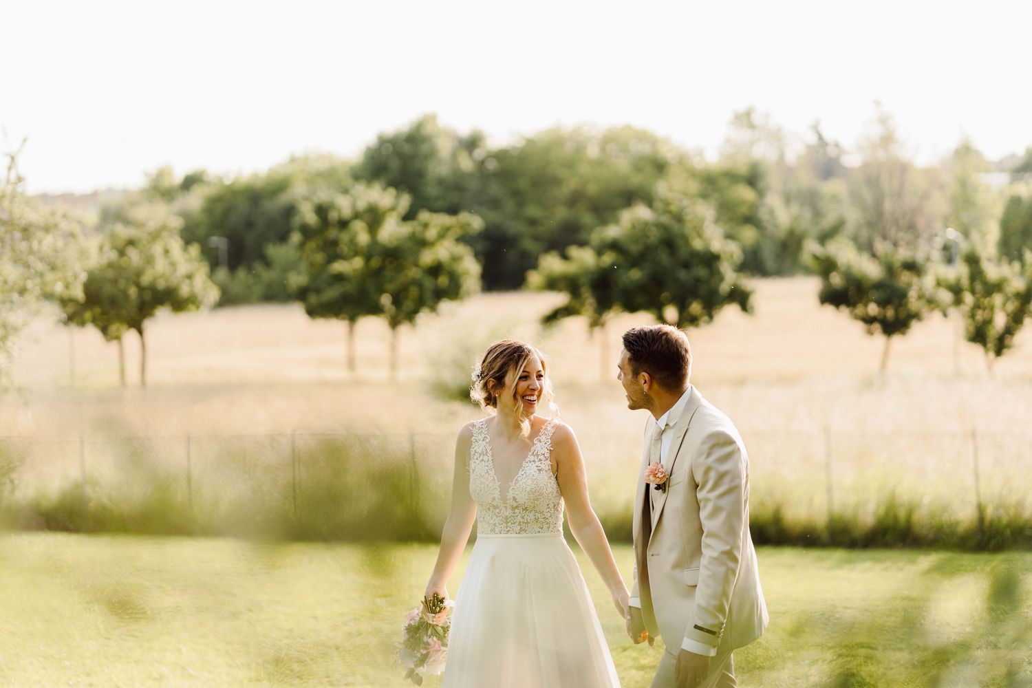 Bride and groom walking hand in hand through a golden sunlit meadow on their wedding day.
